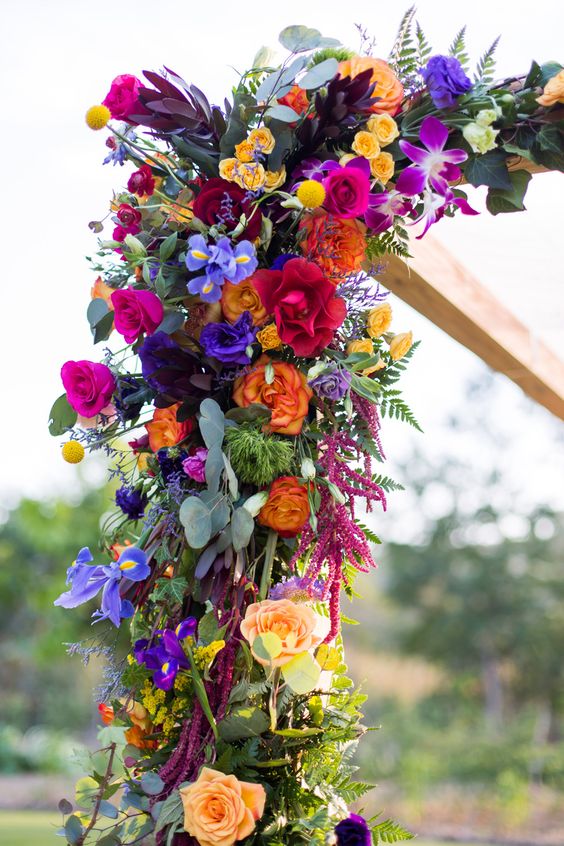a fantastic wedding arch decorated with purple, violet, hot pink, red, orange and yellow blooms and greenery, with much texture