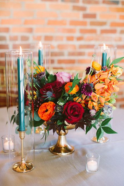 a gorgeous jewel tone wedding centerpiece of orange, yellow, burgundy and fuchsia blooms and some greenery for a fall wedding