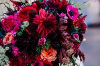 a jewel tone wedding bouquet that includes red, purple, orange, deep purple blooms and greenery and features a sphere shape