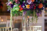 a jewel tone wedding centerpiece of lilac, violet, purple and orange blooms and greenery in a tall vase