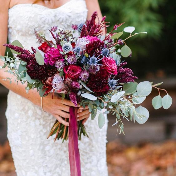 a lovely jewel tone wedding bouquet with hot pink, purple, burgundy blooms, blue thistles and greenery for a fall wedding