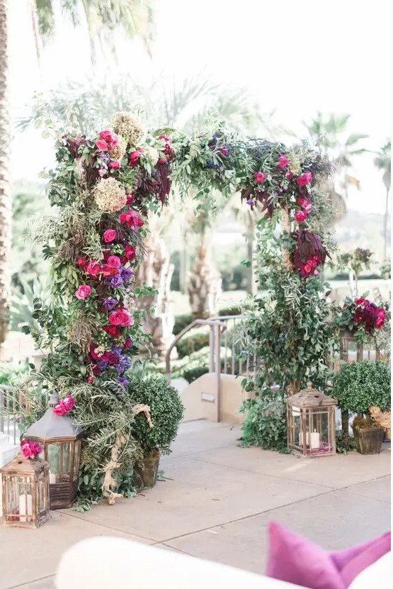 a lush greenery wedidng arch with fuchsia, violet and burgundy blooms and super lush greenery surrounded with candle lanterns and potted plants