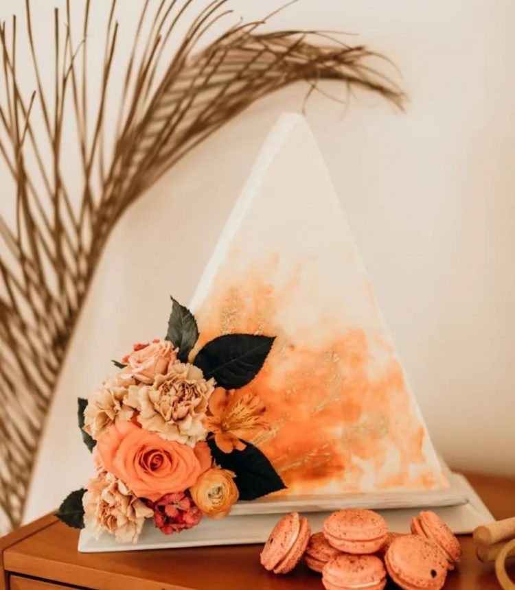 a triangle cake tpo forward wedding cake with orange watercolors, gold glitter and orange and coral blooms and foliage for a bold boho wedding