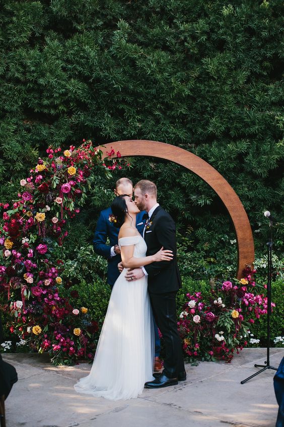 an extra bold jewel tone round wedding arch done with burgundy, depe purple, fuchsia, yellow blooms and greenery
