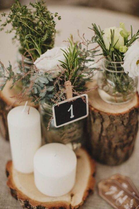 a beautiful cluster wedding centerpiece of tree stumps, white blooms and herbs, a chalkboard number and pillar candles