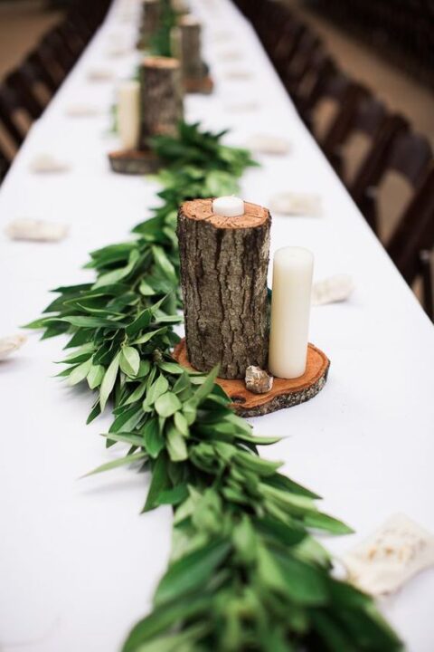 a pretty rustic wedding tablescape with a lush greenery runner, a tree stump on a tree slice, candles and pebbles
