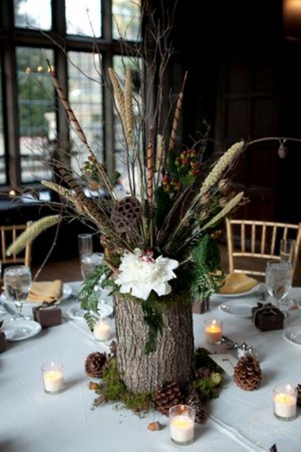 a woodland wedding centerpiece of a moss, pinecones, a tree stump with greenery, blooms, grasses and some candles around is all cool