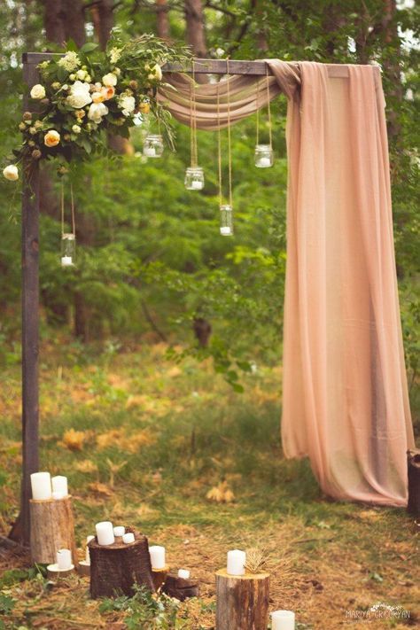 a beautiful rustic wedding arch with blush fabric, hanging candle lanterns, greenery and blooms, tree stumps with candles as additional decor