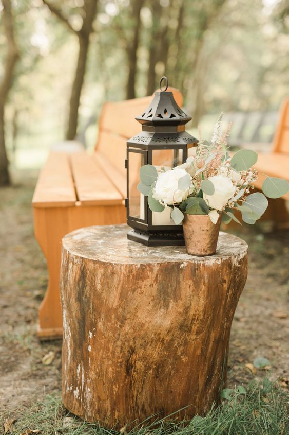 a large tree stump with a candle lantern and a neutral floral arrangement with greenery is a great idea for a rustic wedding