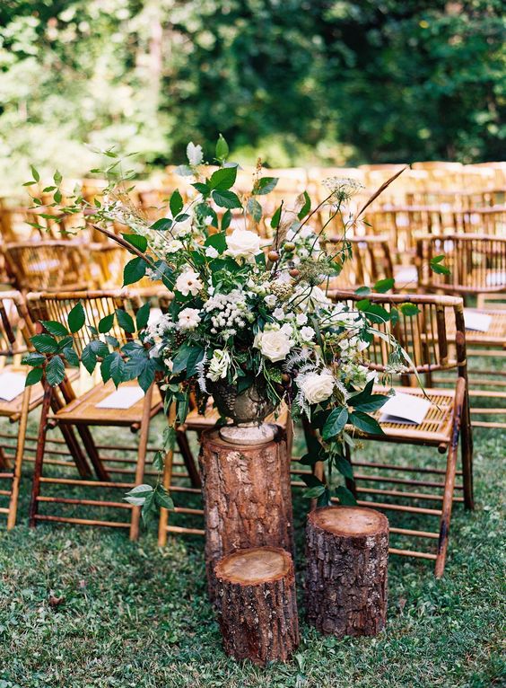 a stylish rustic wedding ceremony space with rattan chairs, tree stumps holding a large white flower arrangement with greenery is wow