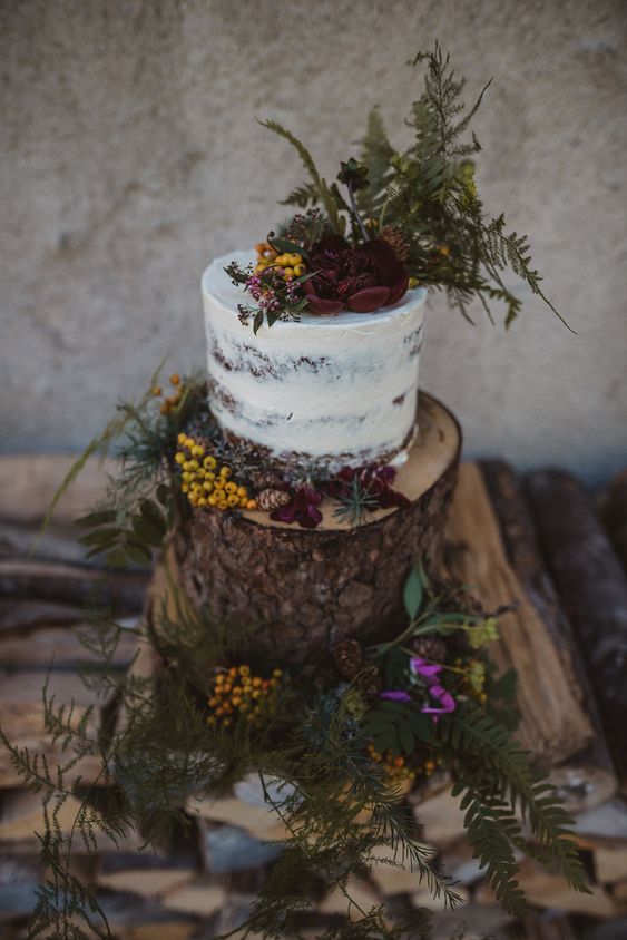 a tree stump decorated with greenery, bold blooms and pinecones, some berries as a stand for a naked wedding cake topped with the same decor