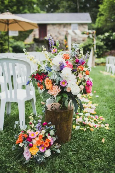 summer wedding aisle decor with a tree stump holding a bright floral arrangement and more arrangements and petals on the ground