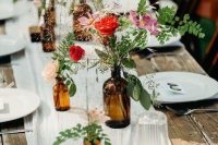 a bright apothecary bottle cluster wedding centerpiece with pink and red blooms, greenery and simple grasses for a relaxed and rustic summer wedding