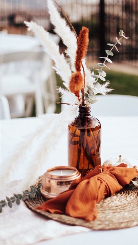 a bright boho wedding centerpiece of an apothecary bottles with greenery and dried grasses, a candle and an orange napkin