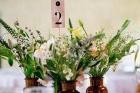 a cluster wedding centerpiece of apothecary bottles, greenery and neutral blooms, a table number for a boho or woodland wedding