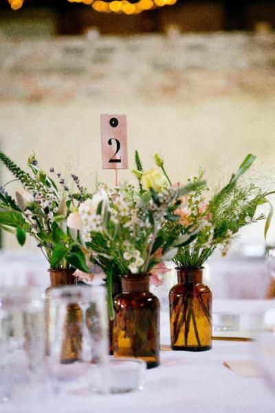 a cluster wedding centerpiece of apothecary bottles, greenery and neutral blooms, a table number for a boho or woodland wedding
