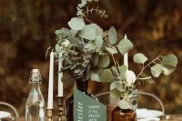 a cluster wedding centerpiece of apothecary bottles, greenery and white blooms, tall and thin candles and a green menu