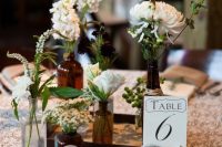 a cluster wedding centerpiece of apothecary bottles with white blooms and greenery and a table number is a nice solution for a rustic wedding