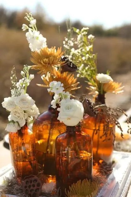 a fall boho wedding centerpiece of apothecary bottles, white and rust blooms and greenery, lotus and dried grasses is cool and relaxed