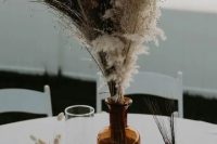 a simple boho fall wedding centerpiece of an apothecary bottle with dried grasses and feathers, candles around is a veyr easy to recreate idea