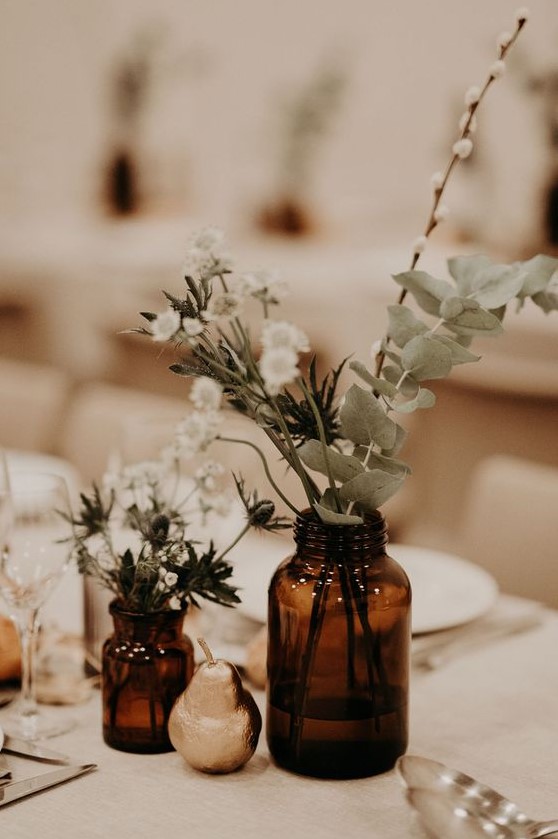 a simple boho wedding centerpiece with apothecary bottles with greenery and white blooms and a gilded pear