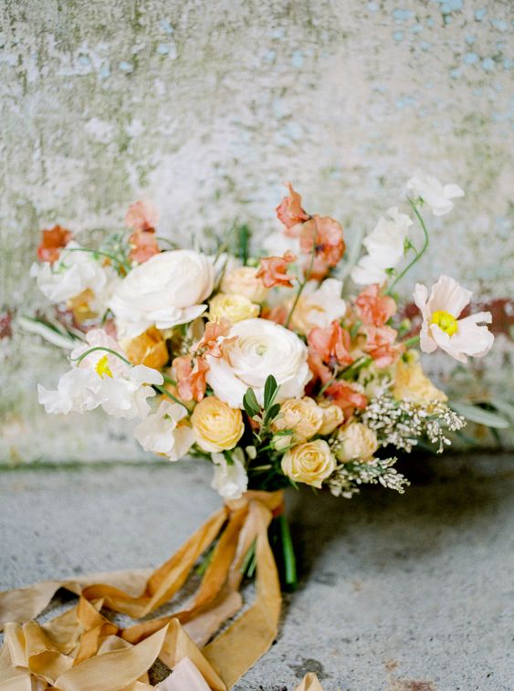 a warm-colored wedding bouquet of white and yellow ranunculus, red and white poppies, greenery and long mustard ribbon is amazing