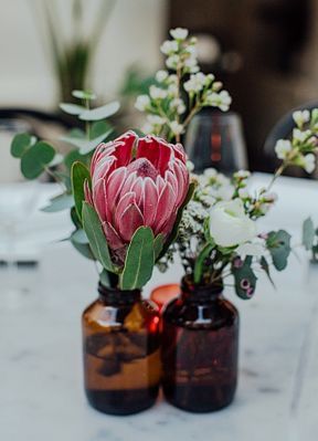 apothecary bottles with white blooms, greenery and a pink king protea are amazing as a creative and bright wedding centerpiece