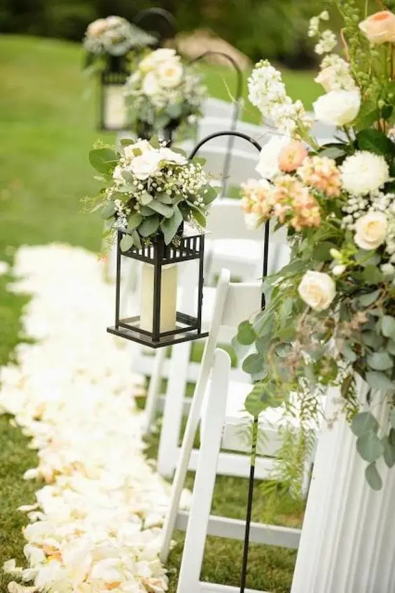 a chic and refined wedding aisle decorated with neutral blooms and greenery, with black candle lanterns topped with blooms and white petals on the ground