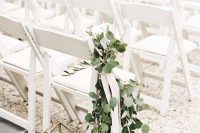 03 a clean and simple wedding ceremony space with white chairs, a chair decorated with greenery and faceted candle lanterns
