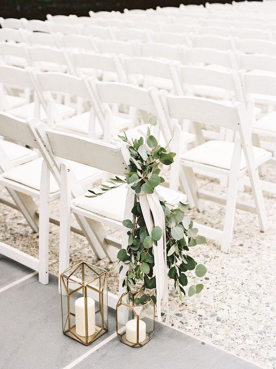 a clean and simple wedding ceremony space with white chairs, a chair decorated with greenery and faceted candle lanterns