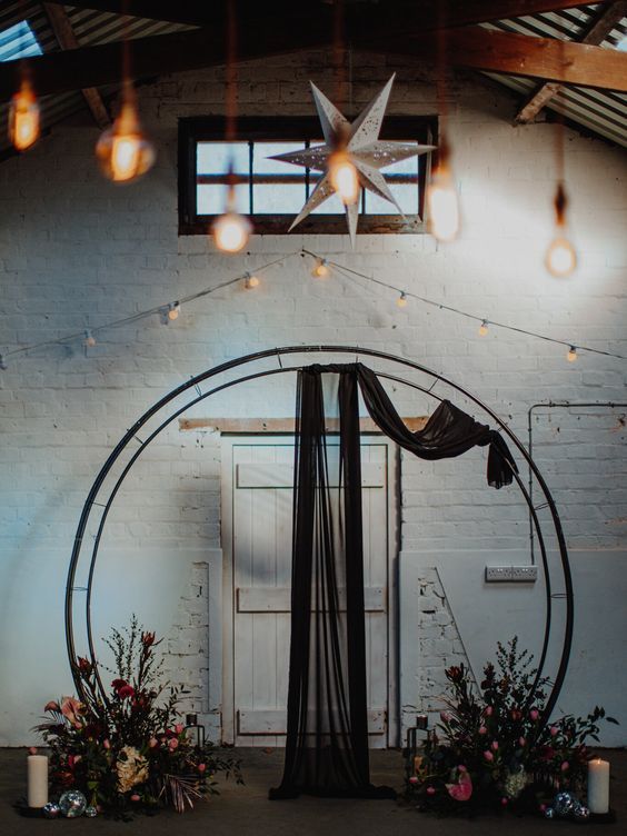 a bold and eye-catchy round wedding arch with bold blooms and dark greenery, with pillar candles and black fabric right on the arch