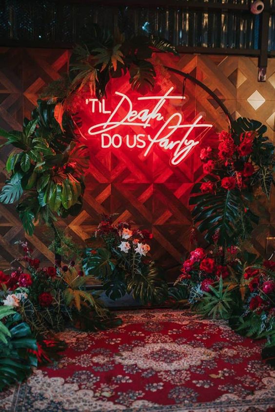 a bold round Halloween wedding arch decorated with fronds and red blooms, a red neon sign is a fantastic idea for such a wedding
