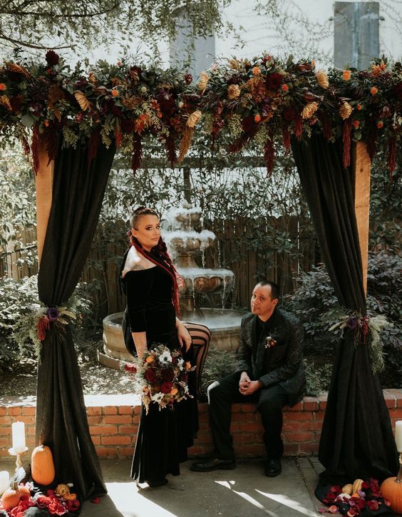 a fantastic moody Halloween wedding arch with black curtains, dark and yellow blooms and greenery, pumpkins, leaves and candles around