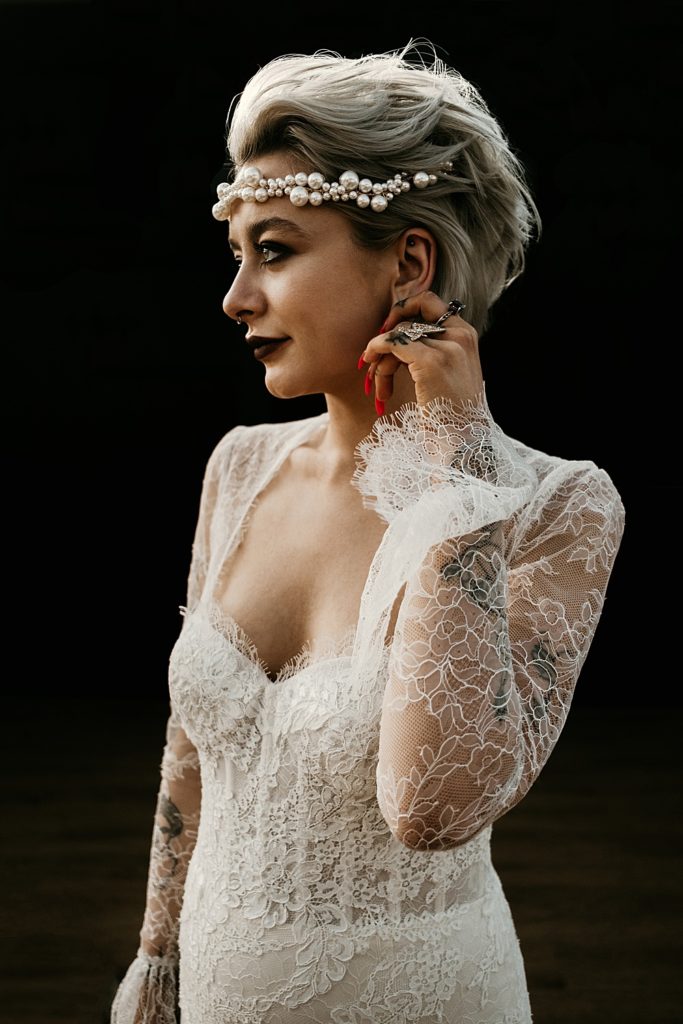 a gorgeous rock n roll bridal look with a romantic lace wedding dress with a deep sweetheart neckline, a statement pearl headpiece, tattoos, statement jewelry and a black lip