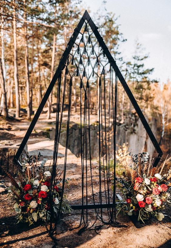 a jaw-dropping black tirangle wedding arch with black macrame, white and red blooms, greenery and dark foliage is wow