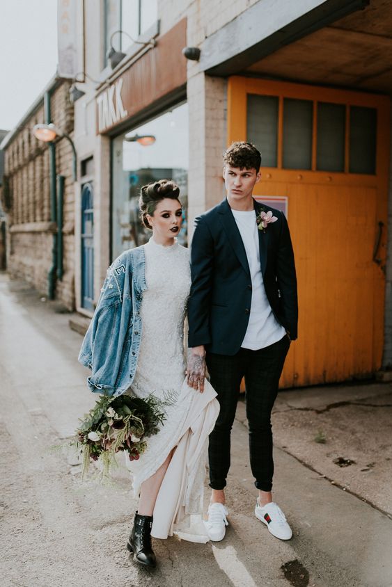 a rock bridal look with a refined and girlish embellished lace wedding dress, black leather boots, an oversized blue denim jacket, a black lip and a catchy hairstyle