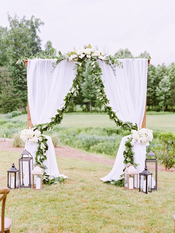 a rustic wedding arch with white curtains, greenery and white blooms and large candle lanterns on both sides is cool