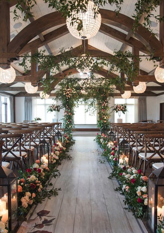 a rustic wedding ceremony space with an aisle lined up with blooms and greenery and large candle lanterns, crystal sphere chandeliers and greenery
