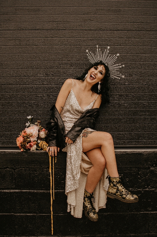 a bold and chic rock n roll bridal look with a white sequin slip wedding dress, a black leather jacket, printed boots, a sunburst headpiece and a chic bouquet