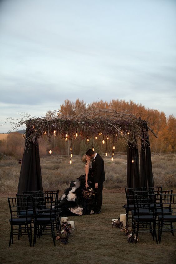 a stylish modern Halloween wedding arch with branches and twigs, bulbs and black curtains is a jaw-dropping idea for a modern Halloween wedding