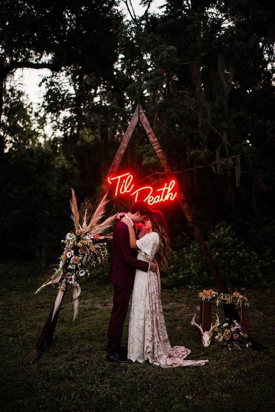 a triangle Halloween wedding arch decorated with neutral and pastel blooms and pampas grass and a red neon sign is amazing