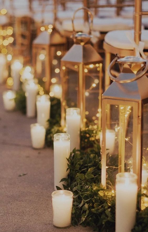 a wedding aisle lined up with giant candle lanterns with candles and LED lights, greenery runners and tall and thin candles