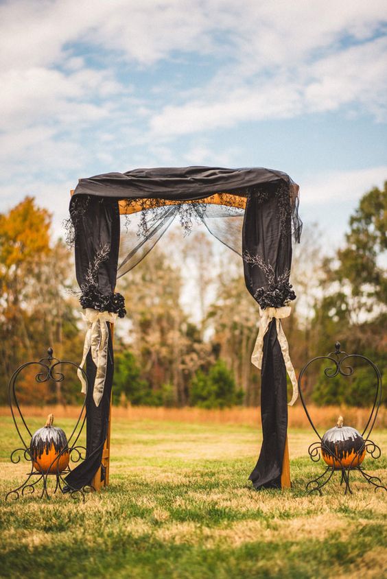 a whimsical Halloween wedding arch with black curtains and black blooms and foliage, white ribbon bows, blackened pumpkins on stands