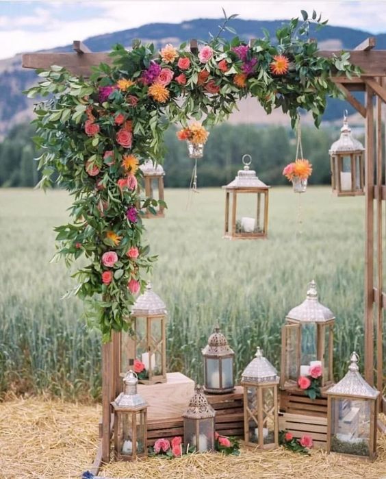 a wedding arch decorated with greenery and bold blooms, crates with wooden candle lanterns and some lanterns hanging down