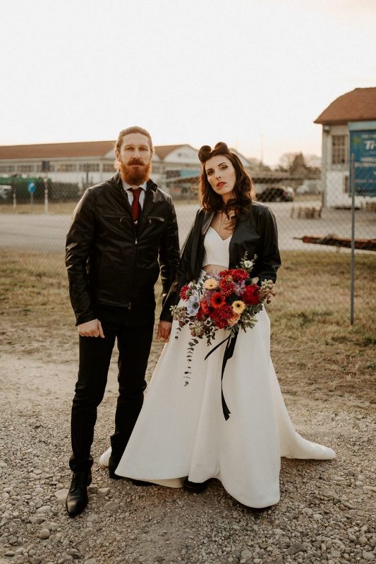 a modern rock n roll bridal look with a crop top and a full skirt with a train, a black leather jacket, a bold hairstyle and a red lip plus a bright bouquet