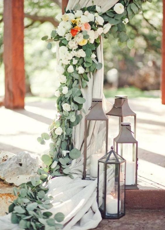 a wedding arch with neutral textiles, greenery and white blooms and large metal candle lanterns around is amazing