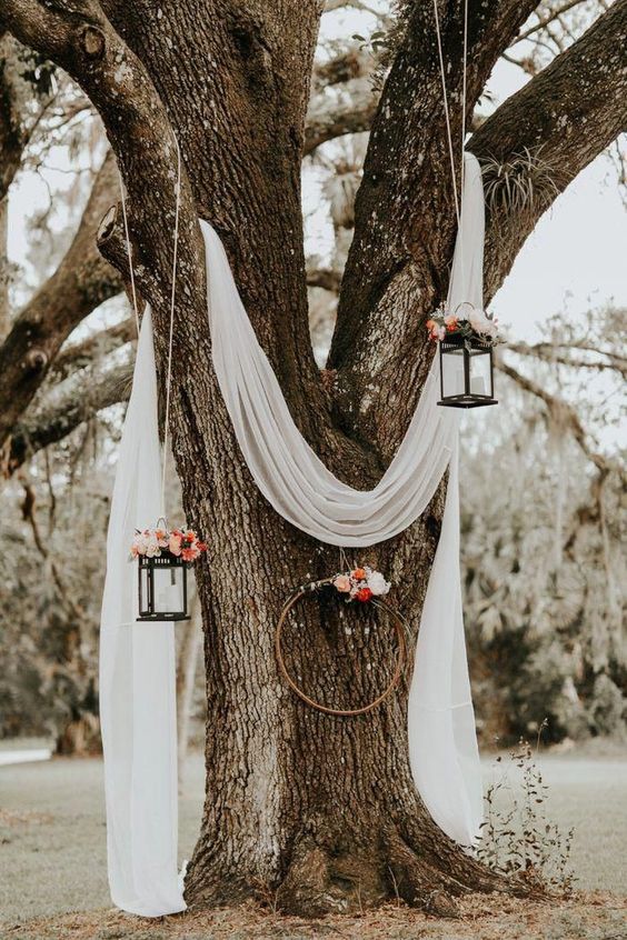 a wedding ceremony space with a living tree, white fabric, hanging candle lanterns and an embroidery hoop decorated with blooms