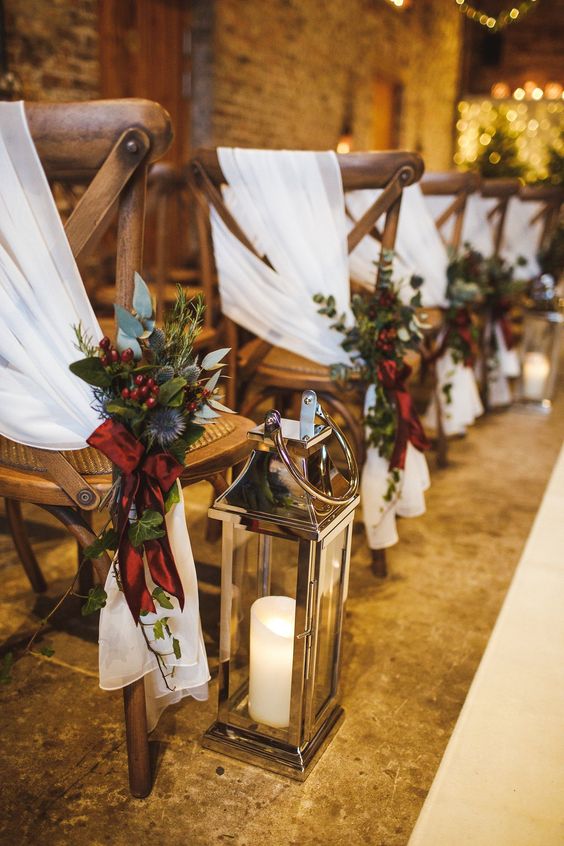 an elegant winter wedding aisle with chairs decorated with white fabric, polished candle lanterns and bright greenery and berry arrangements