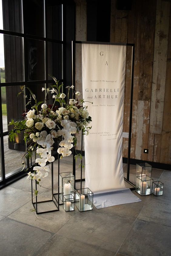 a modern wedding sign on a black frame, with sleek modern candle lanterns, white blooms with greenery on a tall stand is super chic