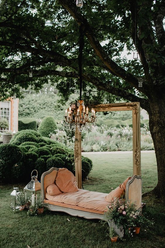 a refined and chic outdoor wedding lounge with a peachy daybed, a frame and a vintage chandelier and lanterns with blooms around